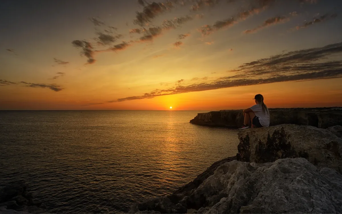 Mulher sentada sobre rochas à beira-mar, olhando o pôr do sol no horizonte