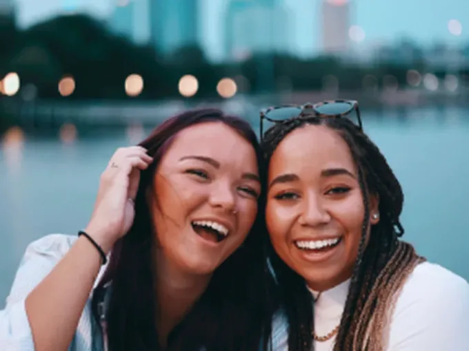 Duas jovens amigas rindo juntas à beira de um lago, com a cidade ao fundo