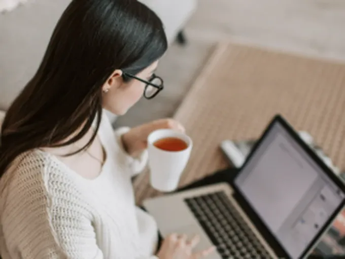Mulher sentada no tapete de casa com uma xícara de chá, em sessão online pelo notebook
